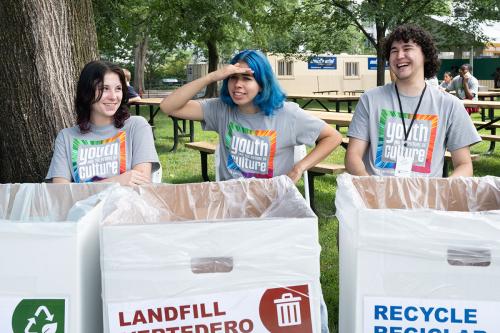 Three young people in matching gray Youth and the Future of Culture T-shirts sit behind cardboard bins marked for compose, landfill, and recycle. The person in the middle, with shoulder-length blue hair, shades her eyes looking into the distance, while the others laugh.