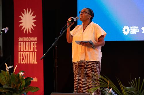 A woman speaks animatedly into a microphone on stage, holding an iPad in one hand. Behind her is a red banner for the Smithsonian Folklife Festival with the Smithsonian sunburst logo.