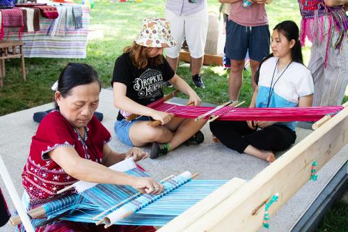 Three people sit cross-legged on a low platform in the grass. Two are using backstrap looms, stretched between the straps around their hips and a wooden crossbeam. One woven textile is bright blue, the other bright red. 