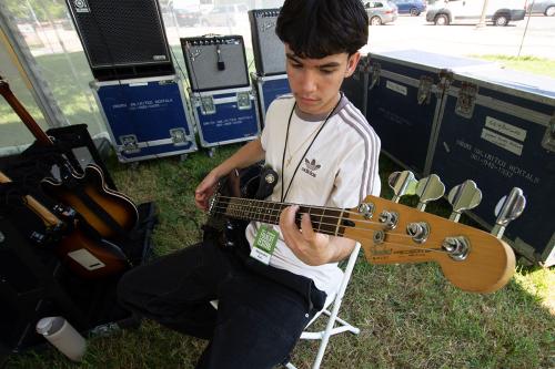 A teen boy wearing a white Adidas shirt sits under a tent surrounded by electric guitars and gear cases, playing an electric bass.