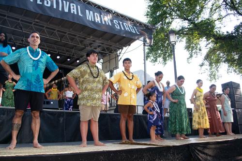 Two line of hula dancers, including adults and children, stand with arms akimbo on two levels of an outdoor stage.