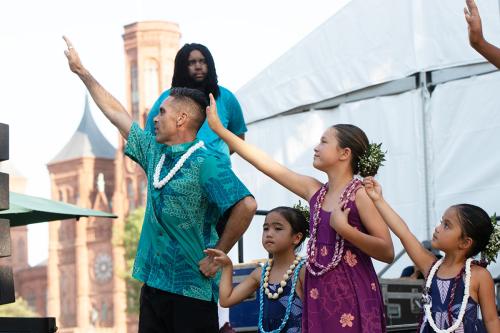 An adult and three children perform hula on stage, each with their right hands raised in unison and wearing leis around their necks. Behind them, an ASL interpreter, a white tent, and the red-brick Smithsonian Castle.
