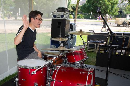 A man sits behind a shiny red drum kit, set up on a low platform in the grass under a tent. He smiles broadly and raises one hand in a wave.