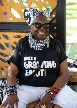 A woman smiles broadly under an outdoor pavilion. She wears a black T-shirt that reads Once a Growing Griot with slits cut in the shoulders, a black-and-white checker kerchief around her neck, a black and white African print crown, and several bangle brac
