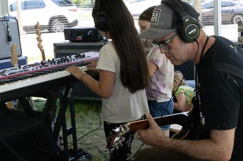 A man wearing a Folkways hat and headphones sits, concentrating as he plays electric guitar. Two young kids behind play an electric keyboard.