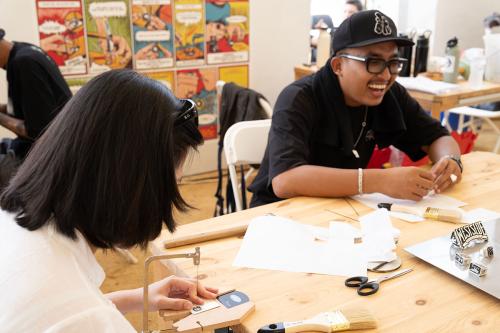 A person with short black hair, seen from behind, is seated at a wooden table and is using a small handsaw to work on a piece of metal jewelry. Across from them, a man wearing a black t-shirt and a black baseball cap is laughing, looking towards the person with the saw.