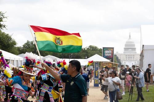 A group of dancers in colorful embroidered outfits and feathered hats dance on a walkway of the National Mall, as a crowd watches and takes photos and a man in the foreground waves a Bolivian flag, with red, yellow, and green stripes. In the distance is t