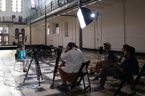 In a large sunlight room, an elder woman sits facing a video camera, a bright studio light, and a group of people sitting, one holding headphones to his ears.