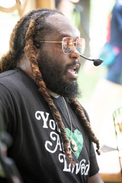 A man sings or speaks on stage, wearing a headset mic and a T-shirt that reads You Are Loved.
