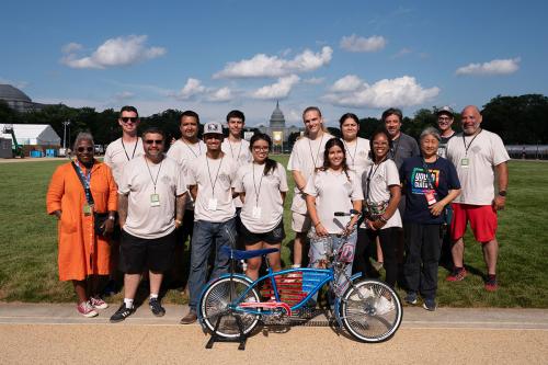 Fourteen people, most in matching white T-shirts, pose on the National Mall, with the Capitol Building in the distance. In front of them is a lowrider bicycle painted blue with an American flag design in the frame, and chrome details.