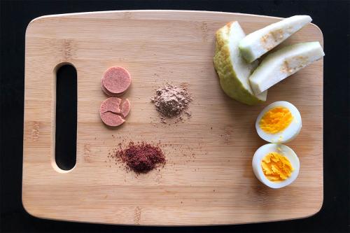 Cutting board containing a lineup of snacks: pink circles, green and white slices of guava, a hard-boiled egg, and two pinched of pinkish powders.