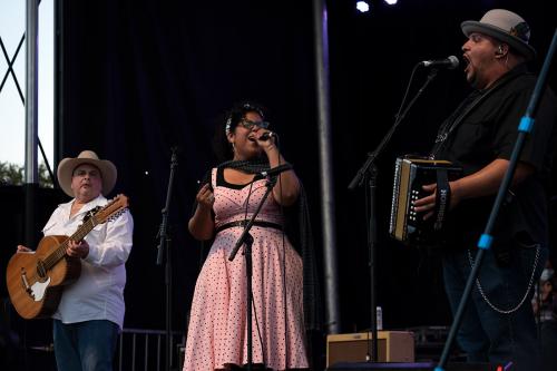 Three people performing on stage: a man with a cowboy hat and a 12-string guitar-type instrument, a woman singing in pink polka dot dress, and a man singing and playing an Hohner accordion.
