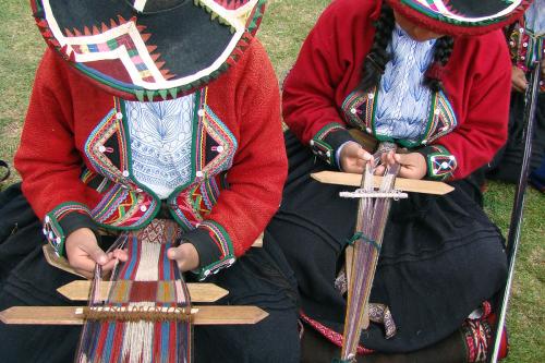 Two people, their faces hidden by wide-brimmed hats, sit on the grass with backstrap looms anchored around their lower backs. In their laps, they use their kinds to guide yarn on the looms.