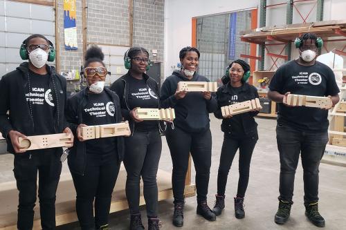 A group of teens in matching black outfits hold up wooden carpentry projects.