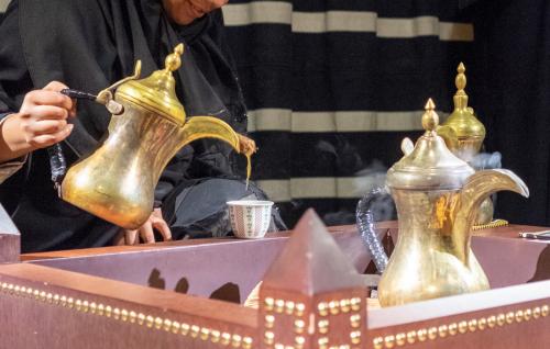 A woman in a black abaya (robe), her face just out of frame, pours coffee from a shiny, golden samovar into a small patterned cup. Two other steaming savors await use.