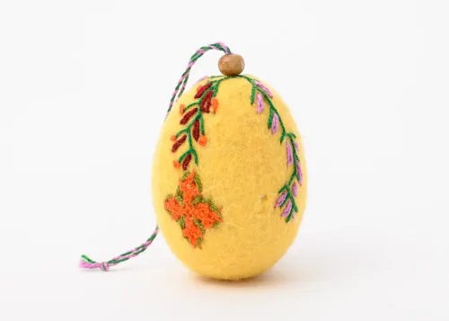 An egg-shaped ornament made of yellow felt is decorated by two colorful strands of floral embroidery that extend midway down the length of the egg. The one on the left terminates in an orange cross.