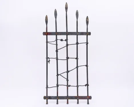A wrought-iron garden gate made of five vertical spires topped with spear-like points is held together by two flat horizonal bars bolted near the top and bottom of the gate. Different lengths of iron rods twist and weave through the spires in an open, asymmetrical pattern.
