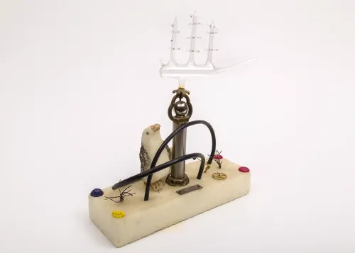 A rectangular base of Styrofoam on which are affixed various objects including a carved Styrofoam penguin, curved wire, watch parts, copper fittings, and a delicate blown-glass ship. 