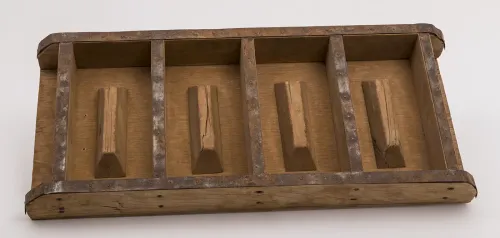 A dark wooden, shallow, rectangular box with no top, divided into four compartments to mold bricks. Rusted metal strips line the top edges.