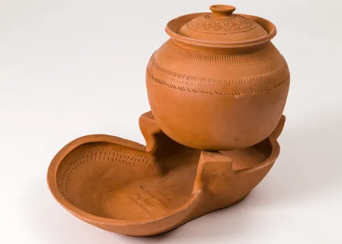 An unglazed ceramic pot with a tight-fitting lid sits on top of a ceramic cookstove formed by two open, rounded, bowl-like sections with raised sides. The back section is taller and has tabs to hold the pot above the adjacent, wider coal bed.