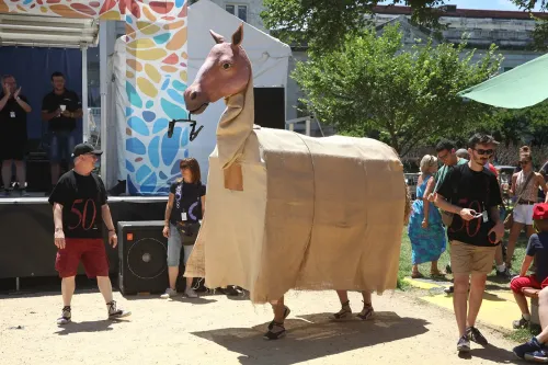 A photograph of a larger-than-life mule “puppet” with a realistic head mounted on a burlap-covered neck and body, as it is carried by two people whose legs are visible beneath the boxy puppet, which is entering a performance space.