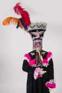 A head to-toe costume consisting of a long, black velvet robe trimmed with white lace and hot-pink marabou feathers, an exaggerated painted face mask, and a large headdress featuring embroidery, a beaded veil, and large colorful ostrich feathers.  