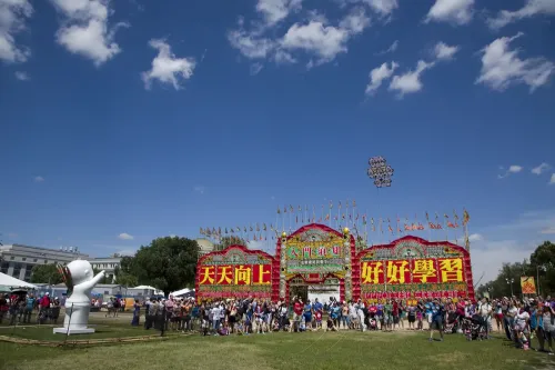 A tall red gateway stretches across the green grass of the National Mall. The gate is decorated with large yellow Chinese characters across the front, and smaller characters in red and white throughout, as well as various flags, banners, and carved and painted designs. “Smithsonian Folklife Festival” is printed above the center section in red lettering.  