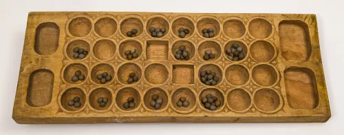 A mancala-like game board of hand-carved wood with four rows of eight shallow cavities each and two oblong cavities on either end. Some hold game pieces made of round, marble-size seeds.