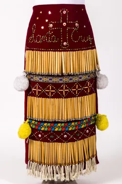 A narrow, red, floor-length cloth skirt intricately decorated with natural river reeds, bells, beadwork, ribbons, and large yarn pom-poms.