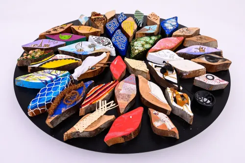 A collection of several dozen colorfully painted cross-sections of palm leaf stems sits on a round black background. The assemblage is photographed from a slight angle, making it appear like a rounded oval, receding into the background, highlighting the dimensionality of the eye-shaped palm paintings.