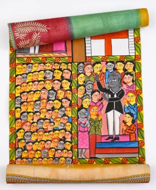 A partially unrolled Indian “story scroll” painting with a multicolored fabric backing features two side-by-side scenes. On the left is a closely packed crowd of faces. On the right, a man holds a microphone and speaks to a close-in group of people surrounding him.