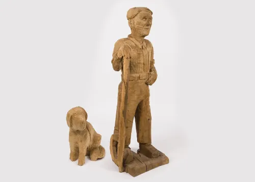 A nearly life-sized wooden figure of a man holding a long-handled, hooked logging tool and standing next to a wooden figure of a seated dog.