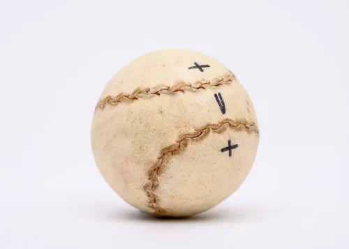 A small, hard leather handball is stitched together with rawhide and marked by three hand-written symbols in black: +, v, +.