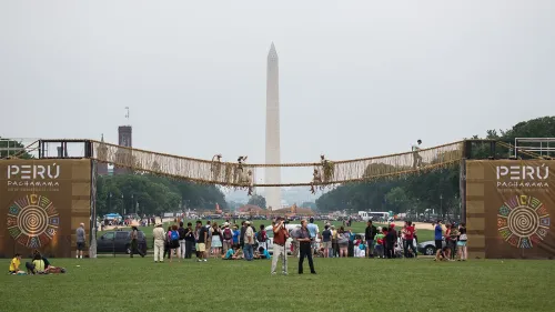A rope bridge spanning across the National Mall is suspended between two towers each featuring the words “Perú: Pachamama” in capital letters. Groups of people are gathered around and under the structure.  