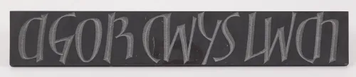 A long rectangular slab of black slate carved with evenly spaced, uppercase, rounded letters that appear light gray and read, in Welsh, “Agor Cyws lwch.”