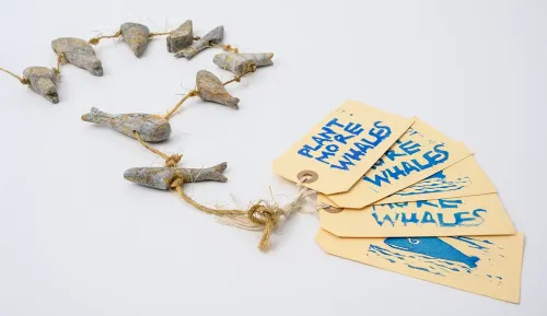 A string of nine small carved stone whales and sharks lies in an S-shaped pattern on a white background. A set of stamped ID tags with the words “Plant more whales” creates a fan at the end.